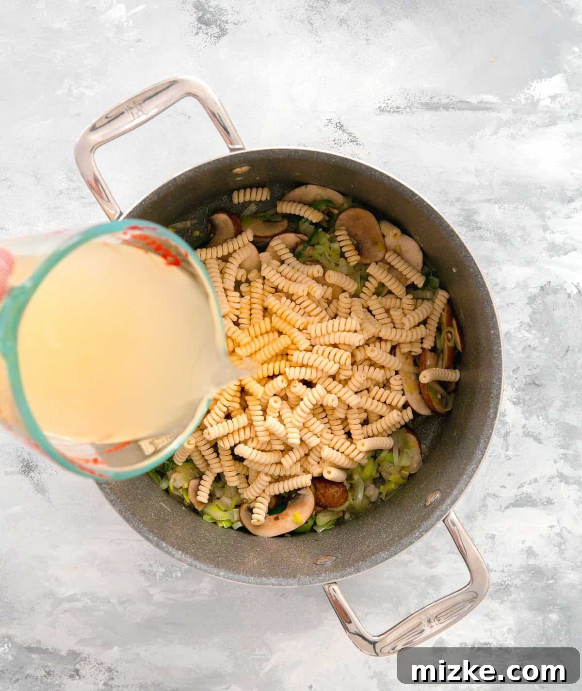 Velvety Mushroom and Leek One-Pot Pasta 9 Chicken stock being poured into the pot over the uncooked pasta and vegetables, ready for simmering.