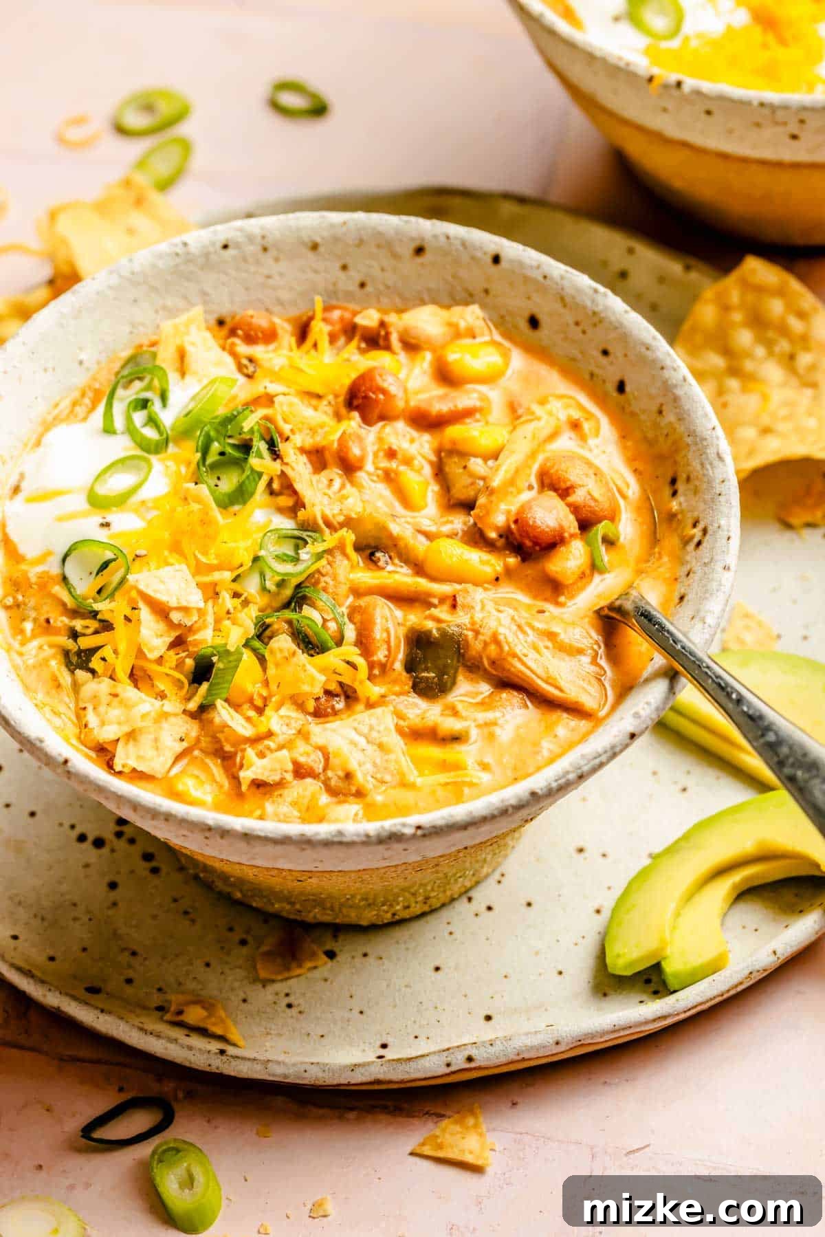 Smoky Southwest Chicken Chili 4 A vibrant bowl of Southwest Chicken Chili with a metal spoon, resting on a white plate alongside fresh avocado slices, ready to be enjoyed.