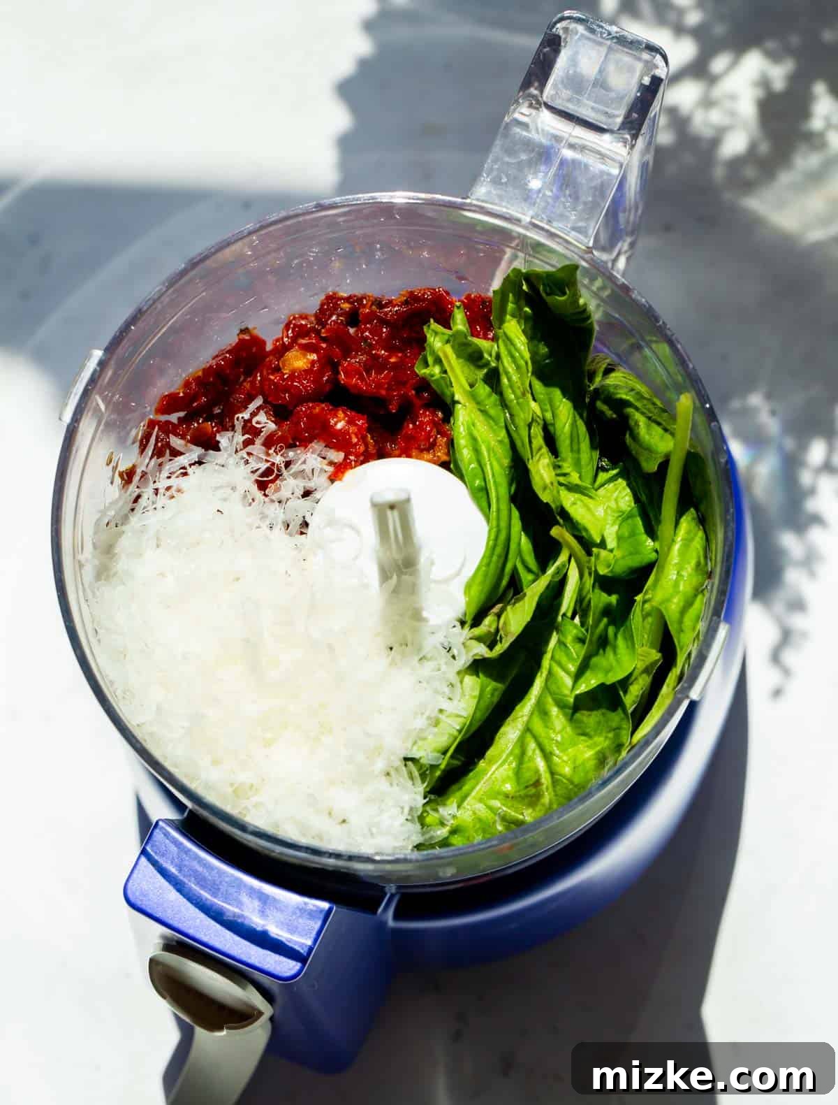 Rich Sun-Dried Tomato Pesto 3 A vibrant collection of fresh basil leaves, rich red sun-dried tomatoes, and finely grated Parmesan cheese, perfectly arranged in the bowl of a food processor, poised for blending.