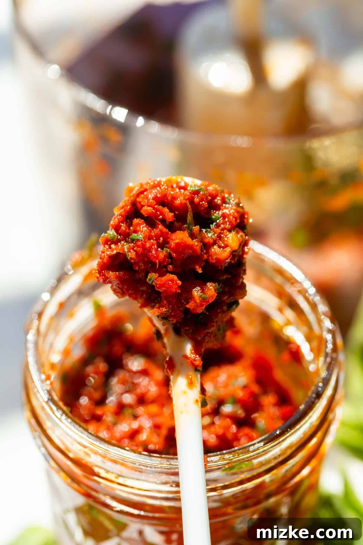 Rich Sun-Dried Tomato Pesto 5 A white spoon delicately lifts a generous dollop of rich red sun-dried tomato pesto from a clear glass jar, showcasing its luscious, thick texture.
