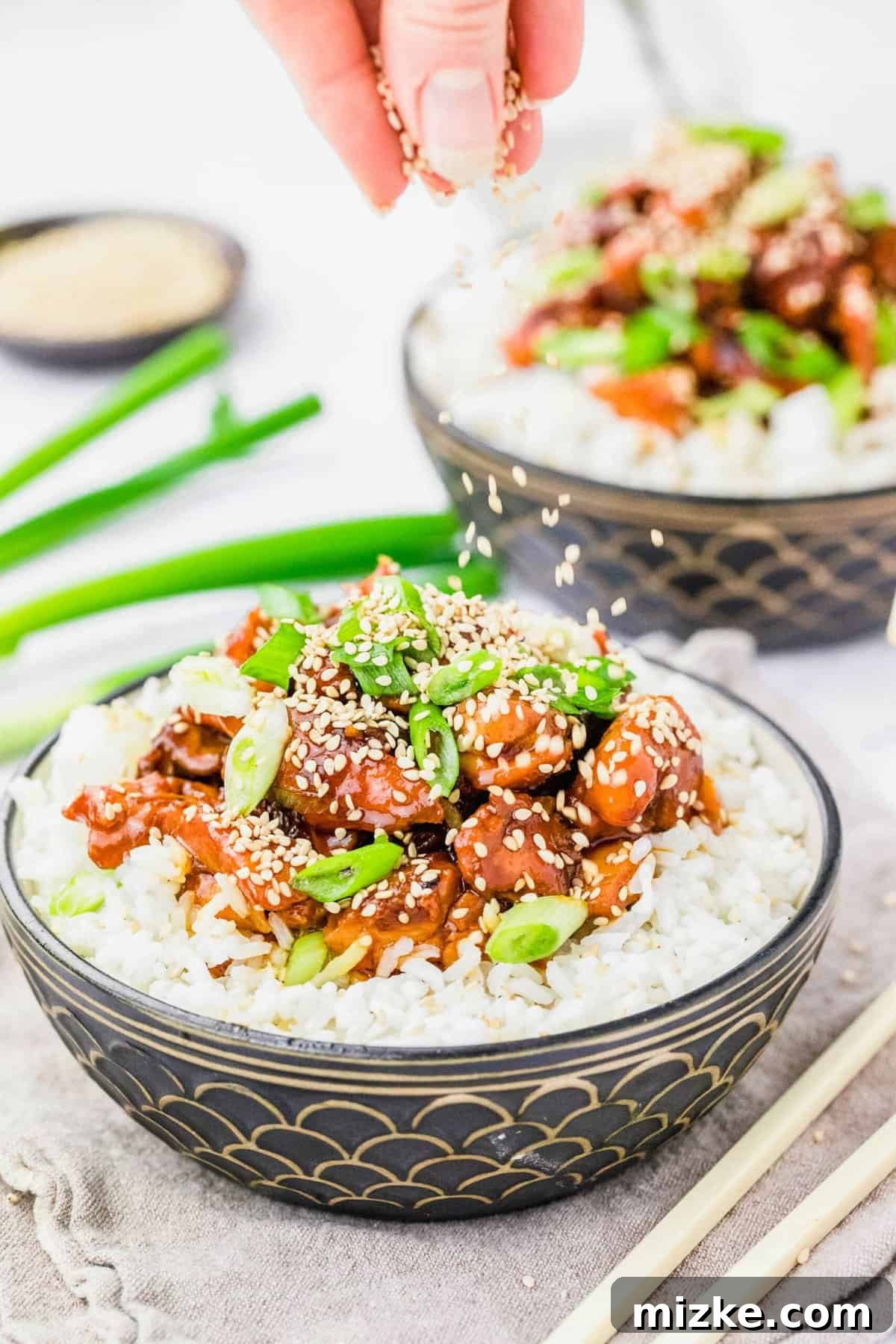 Garnishing Honey Sesame Chicken with Sesame Seeds A hand gently sprinkles white sesame seeds onto a freshly prepared bowl of crockpot honey sesame chicken and rice, adding a final touch of garnish.