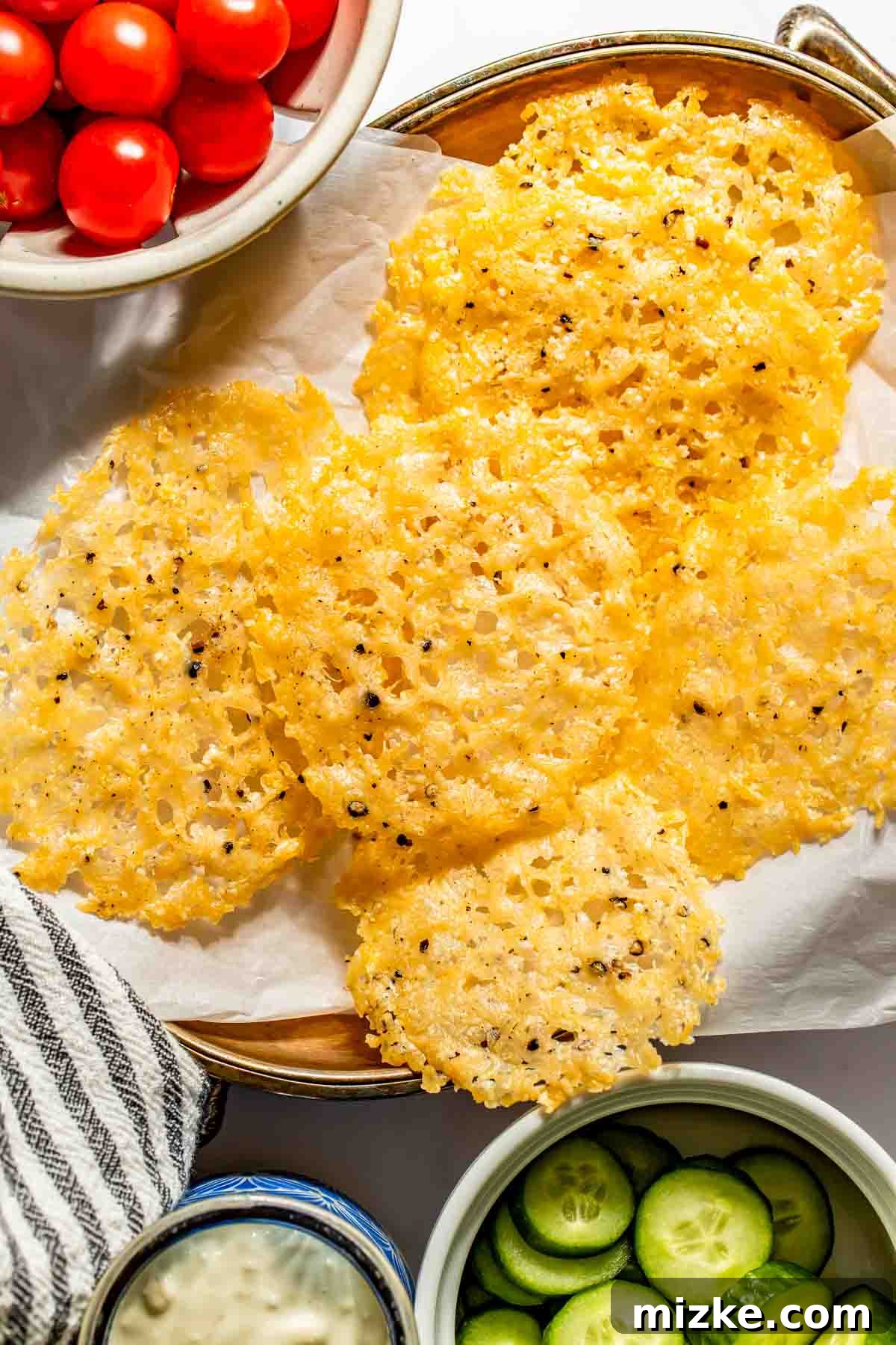 Parmesan Snaps 3 A plate of crispy parmesan cheese crisps on parchment paper, surrounded by bowls of cherry tomatoes, cucumber slices, and a dipping sauce.