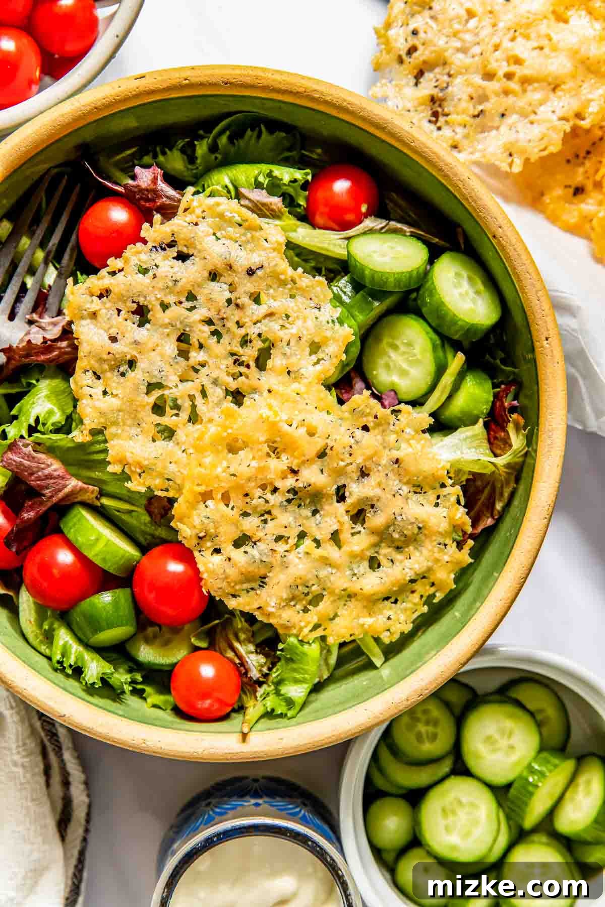 Parmesan Snaps 4 A salad bowl with mixed greens, cherry tomatoes, sliced cucumbers, and topped with two parmesan crisps. Nearby are extra cucumber slices and a small bowl of dressing.