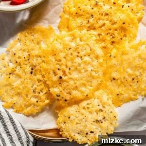 A plate of crispy parmesan cheese crisps on parchment paper, surrounded by bowls of cherry tomatoes, cucumber slices, and a dipping sauce.