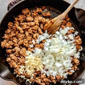 Adding onions and garlic to a skillet full of browned Italian sausage.