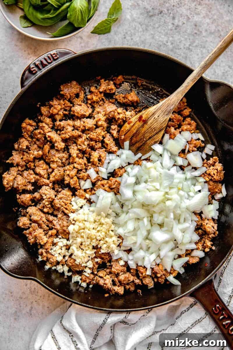 Adding diced onions and minced garlic to the browned Italian sausage in a skillet.