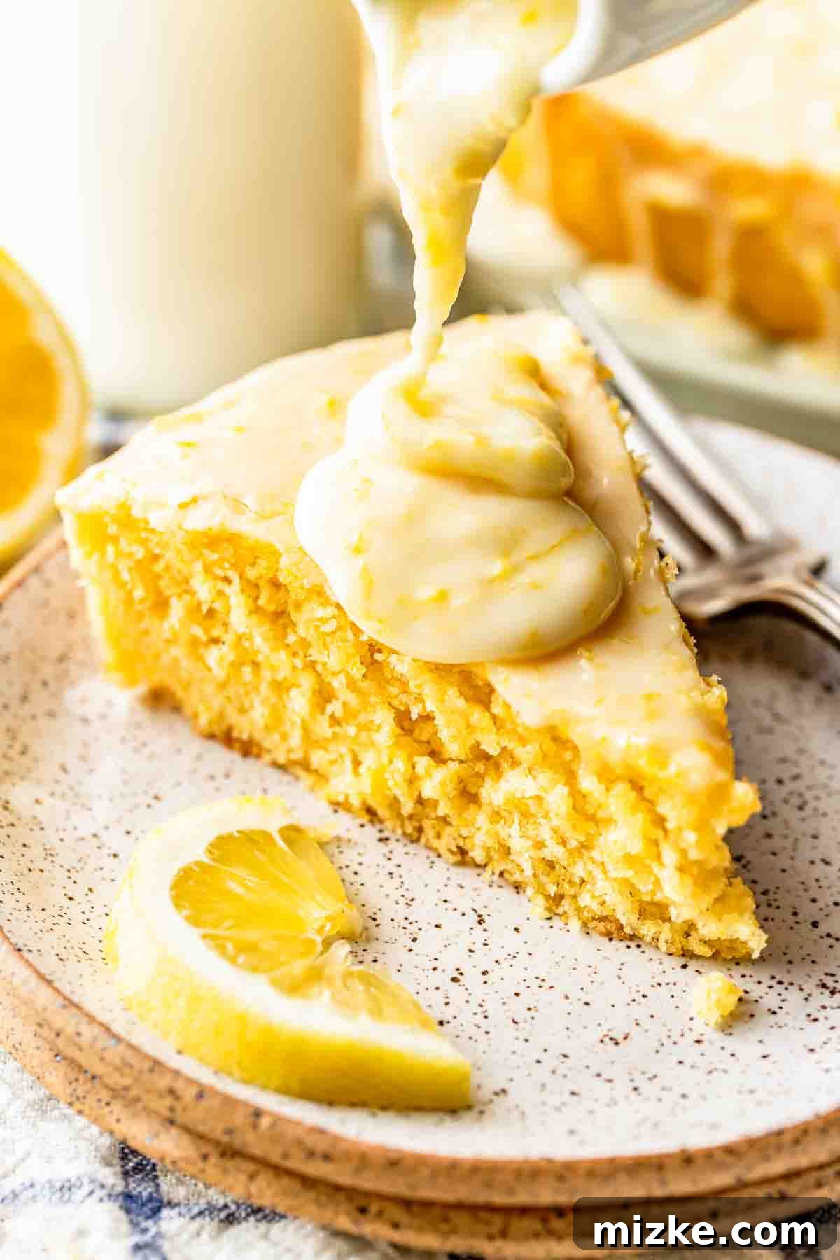 Sunshine Lemon Cornmeal Cake 3 A slice of lemon cake on a plate is topped with thick lemon icing, with a lemon wedge and a fork beside it.
