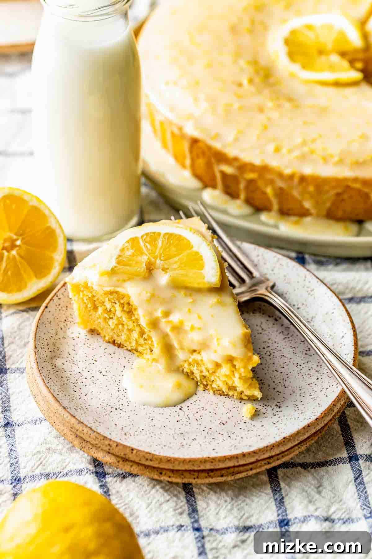 Sunshine Lemon Cornmeal Cake 4 A slice of lemon cake with icing and a lemon slice on top, served on a plate with two forks, next to a bottle of milk and a halved lemon.