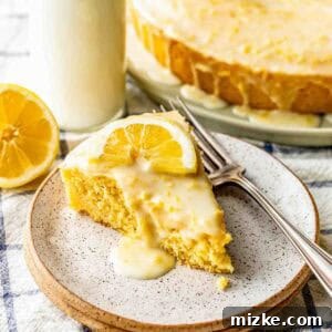 A slice of lemon cake with icing and a lemon slice on top, served on a plate with two forks, next to a bottle of milk and a halved lemon.