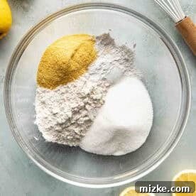 A glass bowl containing flour, granulated sugar, and cornmeal sits on a countertop surrounded by lemons, a sliced lemon, and a metal whisk with a wooden handle.