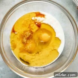 A clear glass bowl containing eggs, milk, vanilla extract, and oil, with a metal whisk and lemon slices nearby on a gray surface.