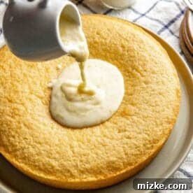 A hand pours lemon glaze from a small pitcher onto a round, lemon cake placed on a plate.