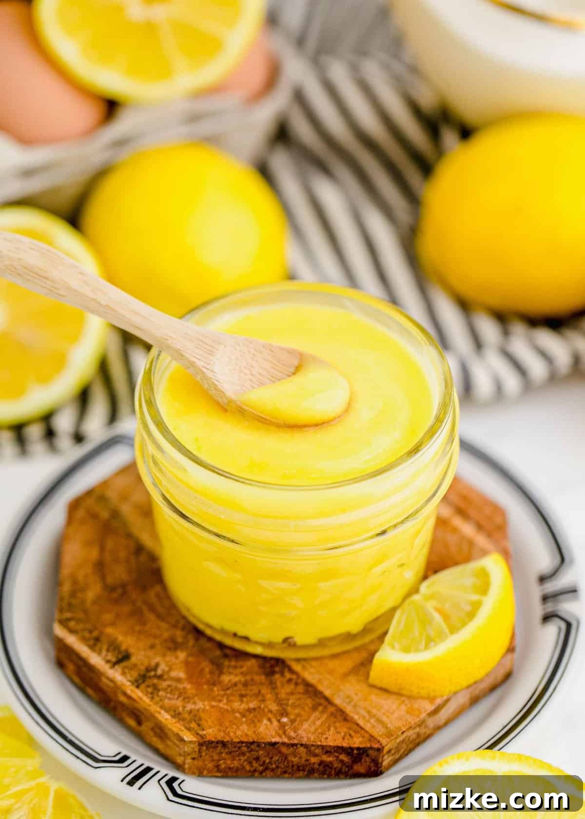 This homemade lemon curd recipe is thick, creamy, and buttery. It's so easy to make and perfect for topping toast, muffins, cakes, and more! A jar of smooth, golden homemade lemon curd with a spoon, accompanied by fresh whole lemons on a rustic surface