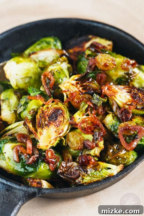 A vibrant side dish of roasted brussels sprouts, beautifully tossed with glistening caramelized shallots and plump golden raisins, all generously topped with crumbly, salty cotija cheese.