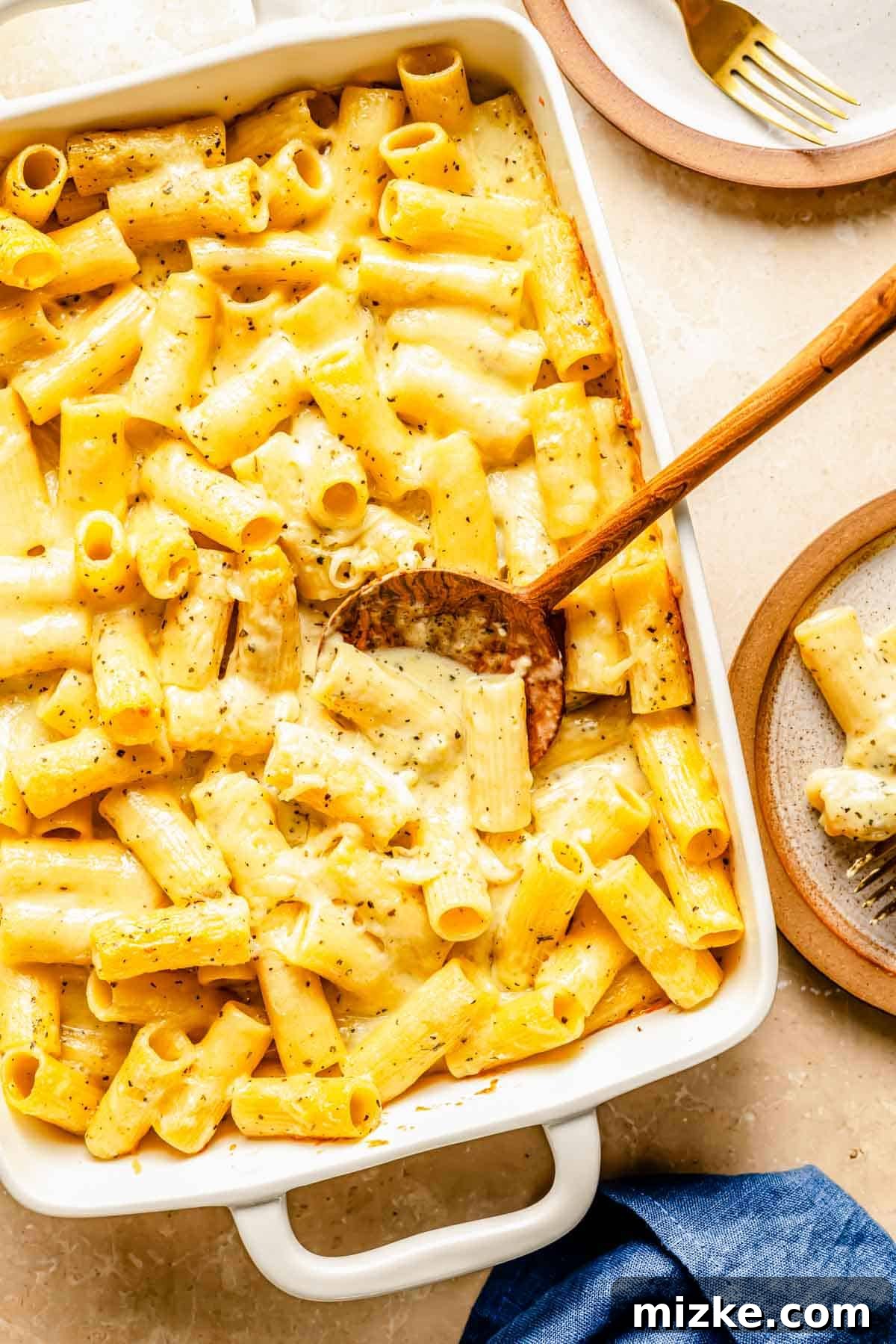 Ultimate Triple Cheese Mac 3 A large white baking dish filled with golden baked three-cheese rigatoni mac and cheese, with a wooden spoon nestled in the center, ready to be served family-style.