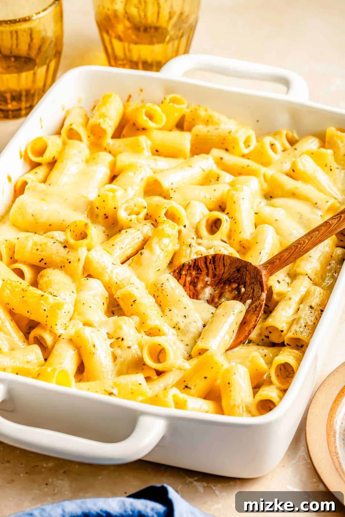 Ultimate Triple Cheese Mac 4 Close-up of baked three cheese rigatoni mac and cheese in a white casserole dish, with a wooden spoon scooping out a portion, highlighting the gooey, melted cheese and perfect golden crust.