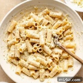 Rigatoni pasta perfectly tossed in the creamy three-cheese sauce mixture in a large white bowl, ensuring every piece is coated.