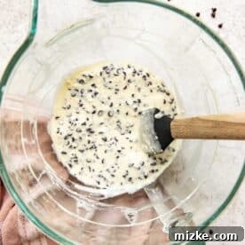 A creamy white mixture with scattered mini chocolate chips in a clear stand mixer bowl, being gently folded with a spatula, ready for the cupcakes.