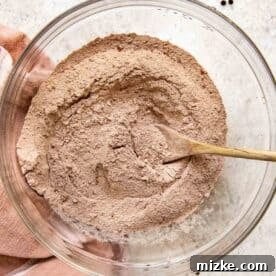 A clear glass bowl filled with dry ingredients including flour and cocoa powder, being mixed with a wooden spoon, showcasing the preparation of the chocolate cake batter.