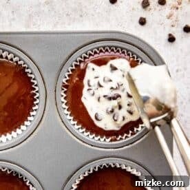 A muffin tin lined with cupcake wrappers, showing chocolate batter in the cups and a spoonful of cream cheese filling being carefully placed into the center of a cupcake.