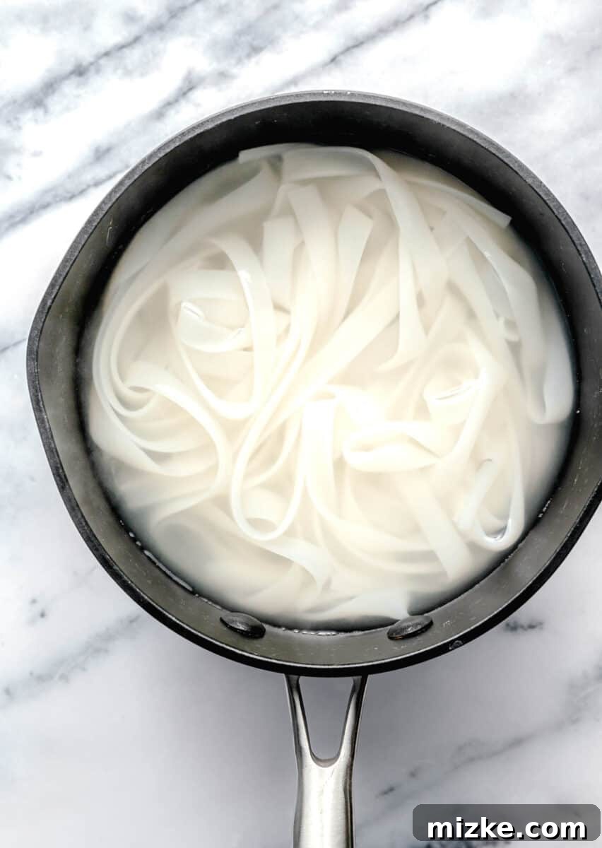 Wide rice noodles soaking in a pot of hot water, preparing for the stir-fry.