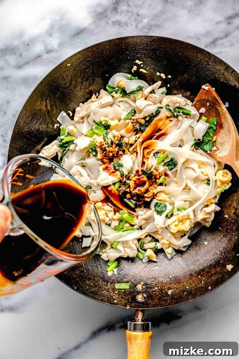 The prepared Pad See Ew sauce being poured into a wok containing rice noodles, scrambled eggs, and Chinese broccoli, ready to be mixed.