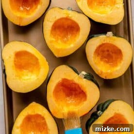 Halved acorn squash on a baking sheet with a brush applying oil