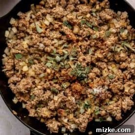 Ground sausage and diced onions cooking in a dark green cast iron skillet, garnished with fresh chopped sage.