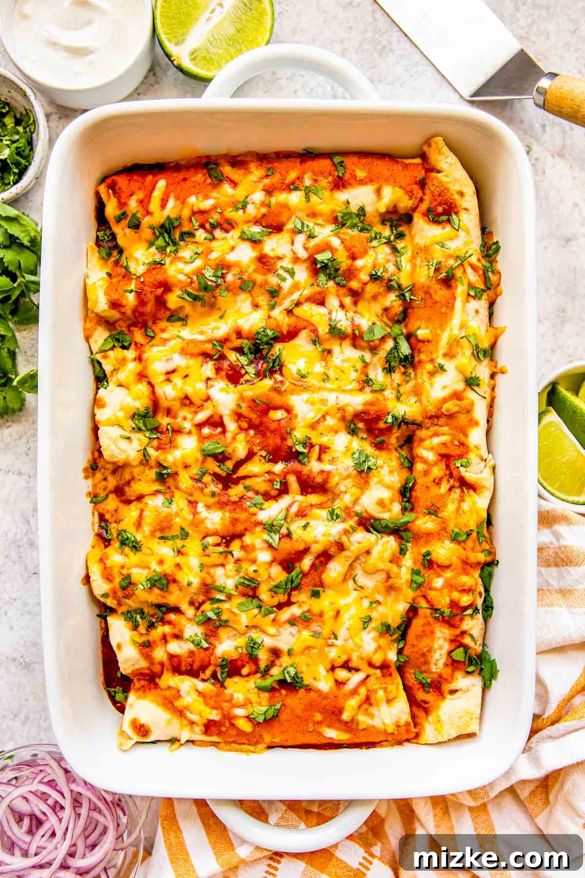 Effortless Homemade Chicken Enchiladas 2 Golden-brown chicken enchiladas nestled in a white baking dish, garnished with fresh cilantro, alongside vibrant lime wedges, creamy sour cream, and thinly sliced red onions, ready to be served for a delightful Mexican meal.