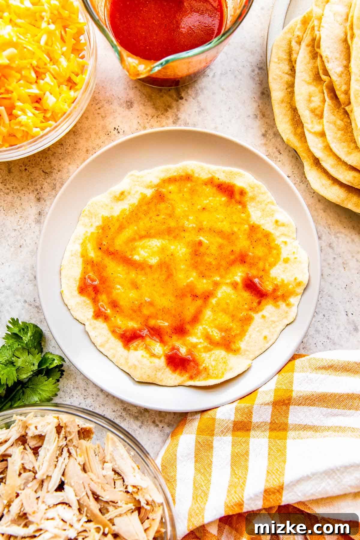 Effortless Homemade Chicken Enchiladas 4 A flour tortilla lies flat on a plate, with a tablespoon of vibrant red enchilada sauce spread evenly across its surface, ready for filling.