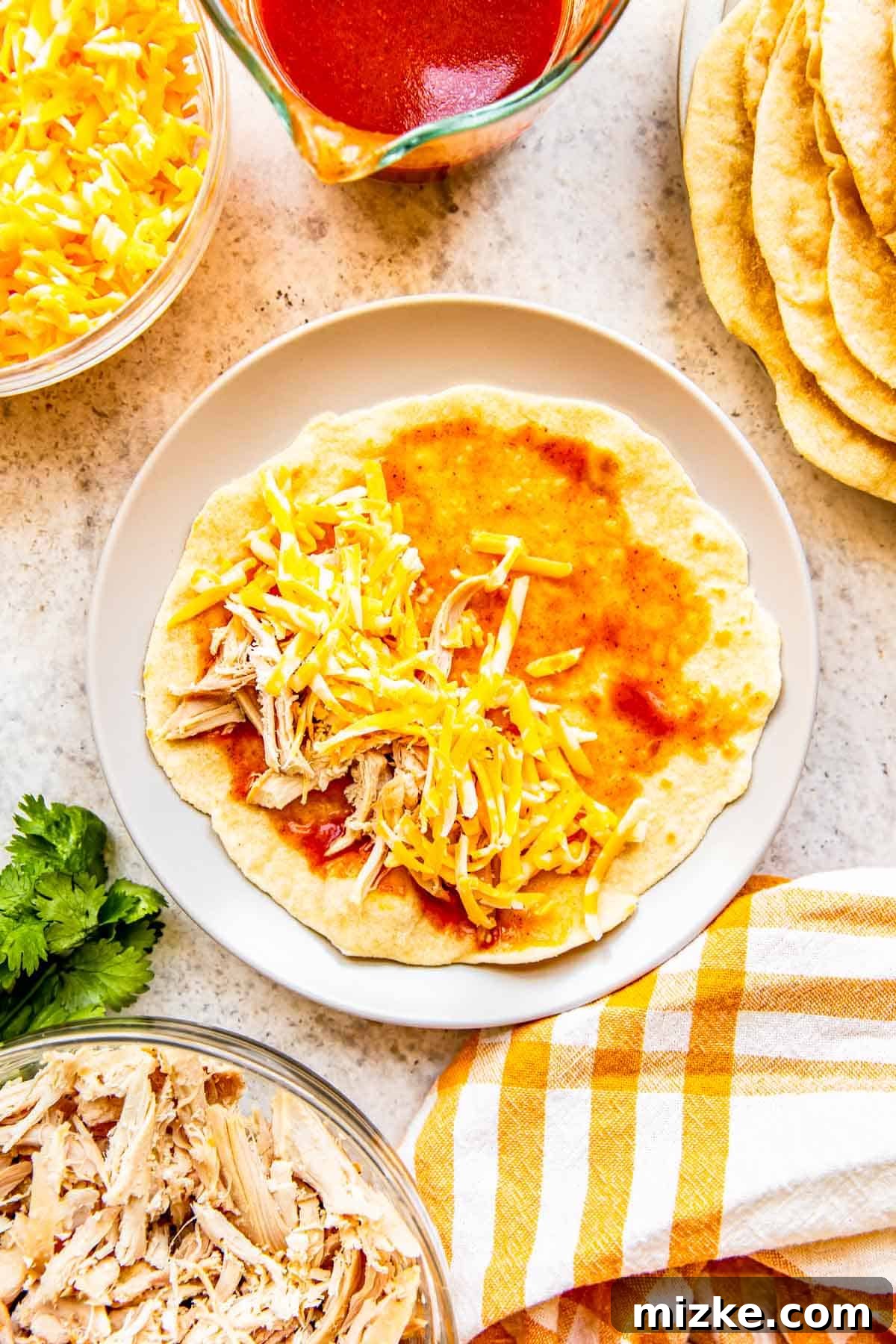 Effortless Homemade Chicken Enchiladas 5 Shredded rotisserie chicken and a generous sprinkle of grated Colby Jack cheese are carefully placed on a flour tortilla already coated with red enchilada sauce, preparing for rolling.