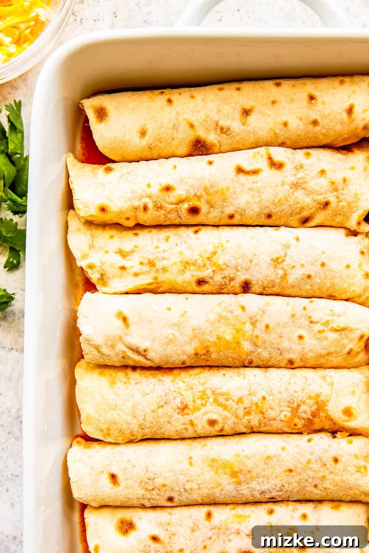 Effortless Homemade Chicken Enchiladas 6 Neatly rolled chicken enchiladas are perfectly aligned seam-side down in a white ceramic baking dish, ready for the final touches.