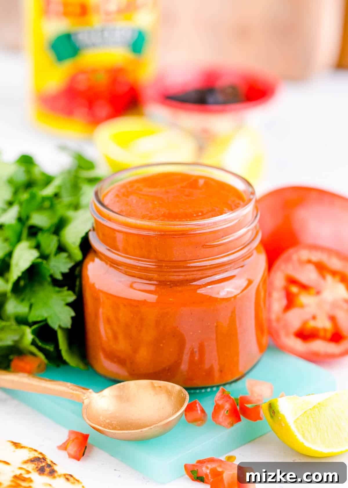 Easy Blender Red Enchilada Sauce 2 Vibrant red enchilada sauce in a clear glass jar, resting on a blue cutting board, surrounded by fresh ingredients like tomatoes, onions, and chili