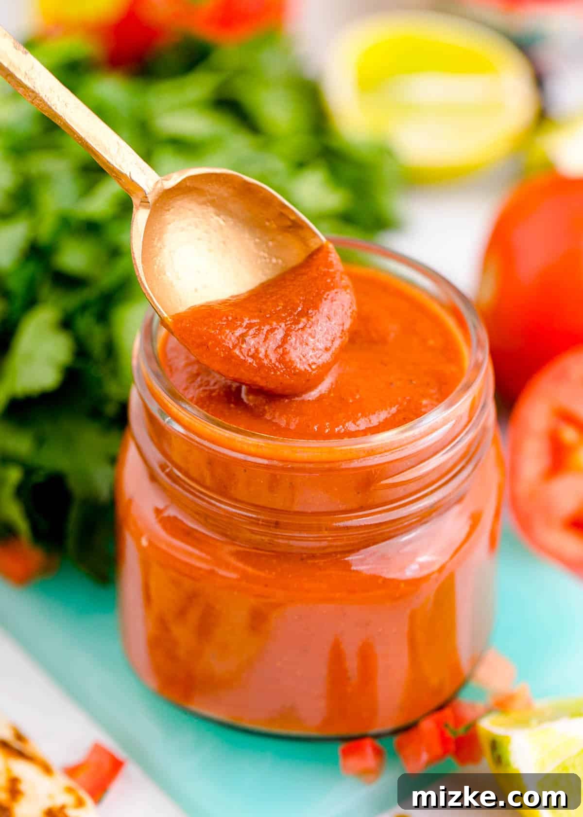 Easy Blender Red Enchilada Sauce 3 A spoon delicately dips into a glass jar of homemade red enchilada sauce, showcasing its smooth texture and rich color on a blue cutting board