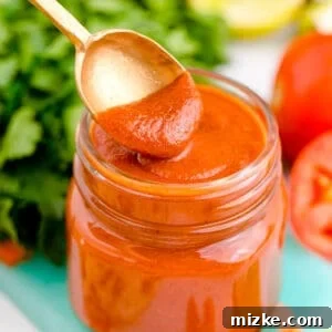 A spoon dips into red enchilada sauce in a glass jar on top of a blue cutting board