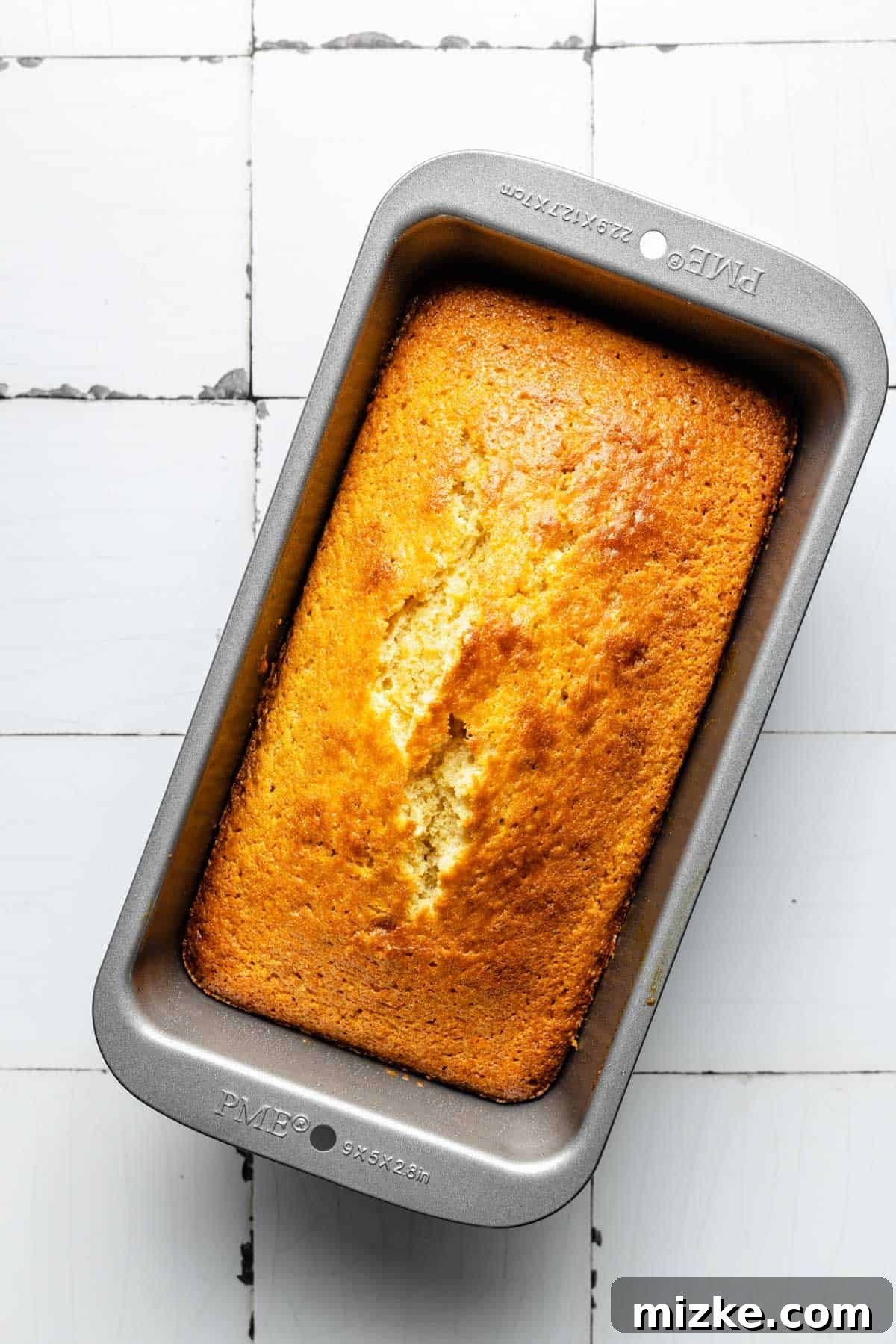 A golden-brown, freshly baked pound cake sits in a loaf pan, cooling slightly before being removed.