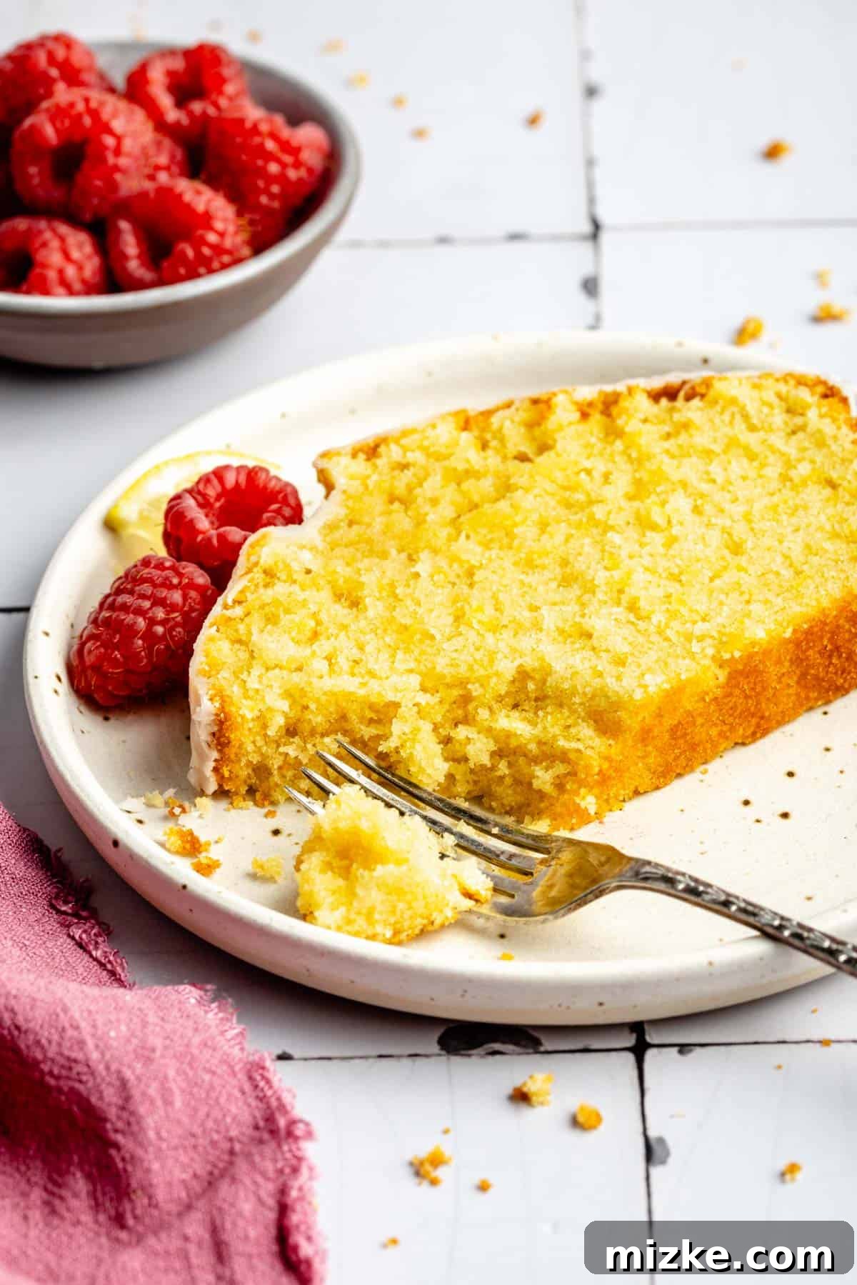 A delicate fork rests on a white plate beside a single, perfectly sliced piece of lemon pound cake, ready to be enjoyed.