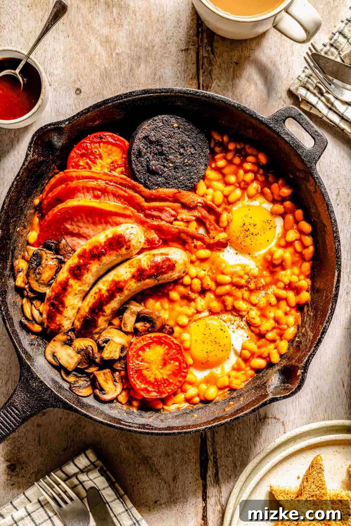 A cast iron skillet filled with a traditional Full English Breakfast, including baked beans, fried eggs, sausages, back bacon, black pudding, mushrooms, and grilled tomatoes, all arranged on a rustic wooden table.