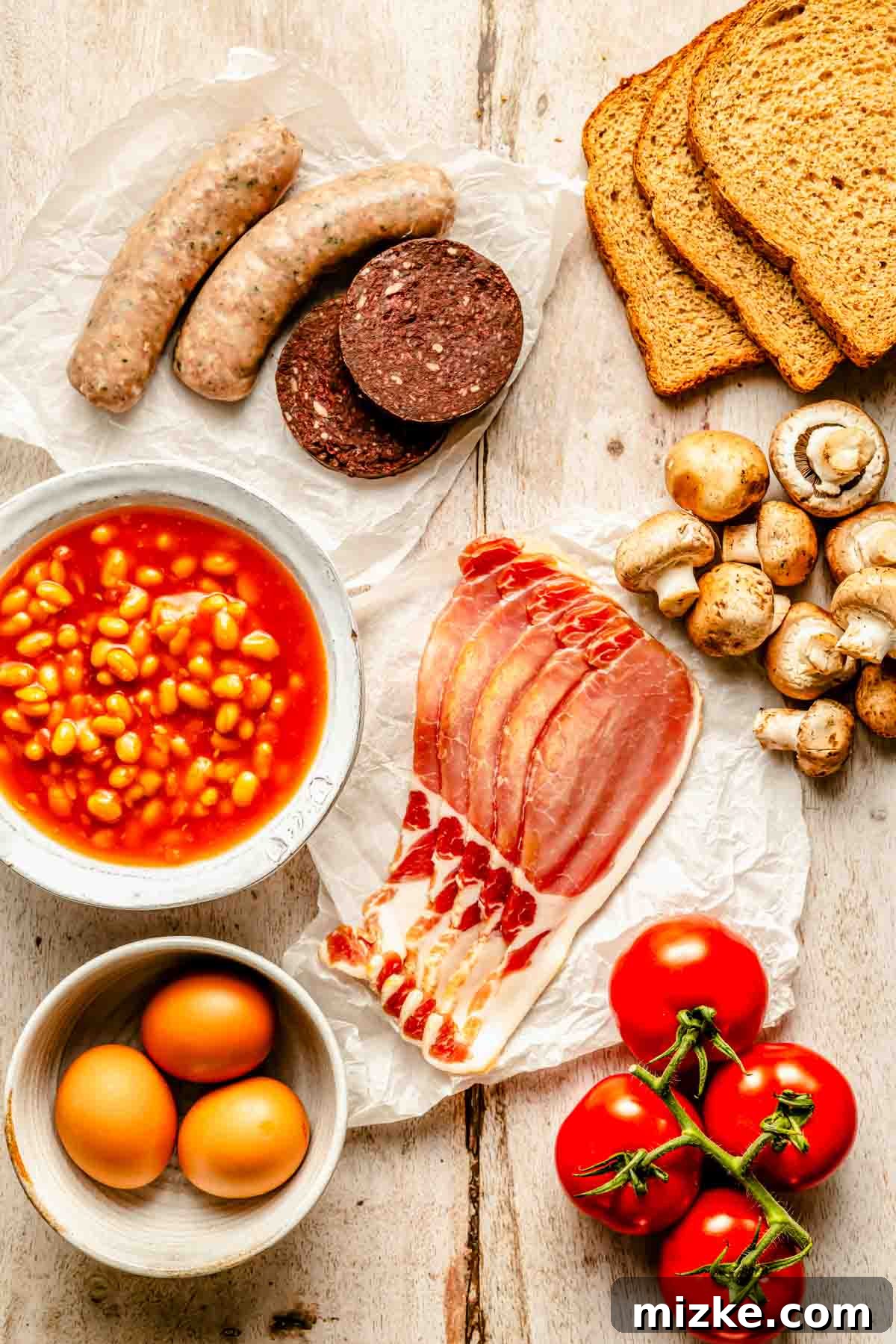 All the fresh ingredients meticulously laid out for a Full English Breakfast, including plump sausages, rich black pudding, a can of baked beans, streaky back bacon, farm-fresh eggs, ripe tomatoes, savory mushrooms, and slices of artisan bread, all arranged invitingly on a wooden surface.