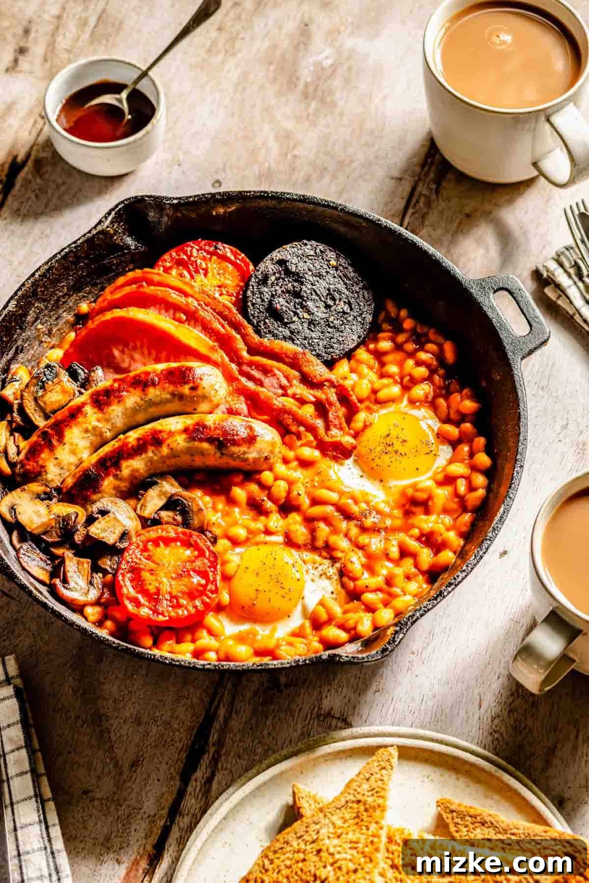 A beautifully arranged cast iron skillet showcasing a complete Full English Breakfast: perfectly fried eggs atop baked beans, alongside sizzling sausages, crispy back bacon, sliced tomatoes, savory mushrooms, and a piece of black pudding. Toasted bread and steaming cups of tea are artfully placed on the side, ready for enjoyment.