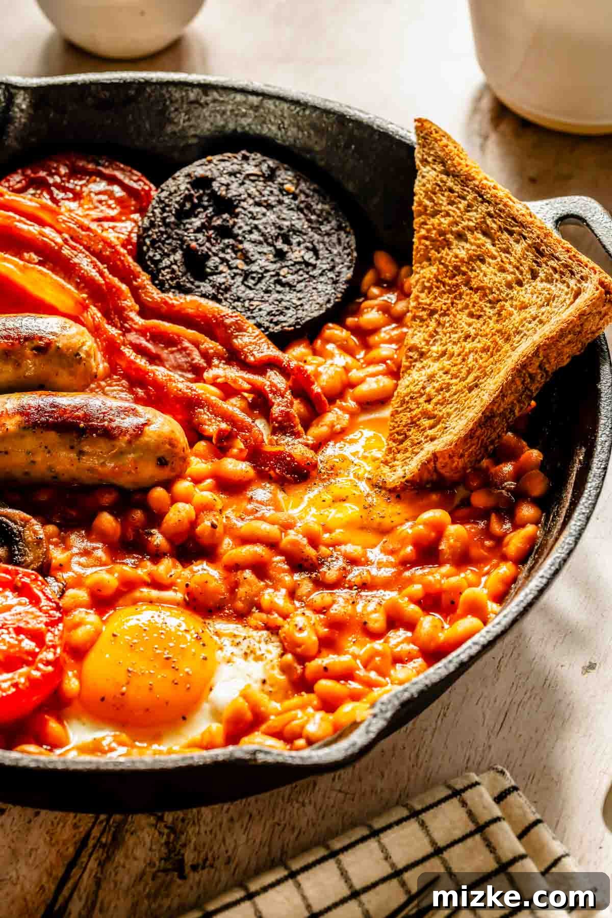 A close-up view of a single-serving Full English Breakfast presented in a cast iron skillet. It features perfectly cooked baked beans, a fried egg with a runny yolk, two savory sausages, two slices of back bacon, a piece of black pudding, a grilled tomato half, and a crisp slice of toast, all resting on a wooden table.