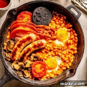 A cast iron skillet with baked beans, fried eggs, sausages, back bacon, black pudding, mushrooms, and grilled tomatoes on a wooden table.