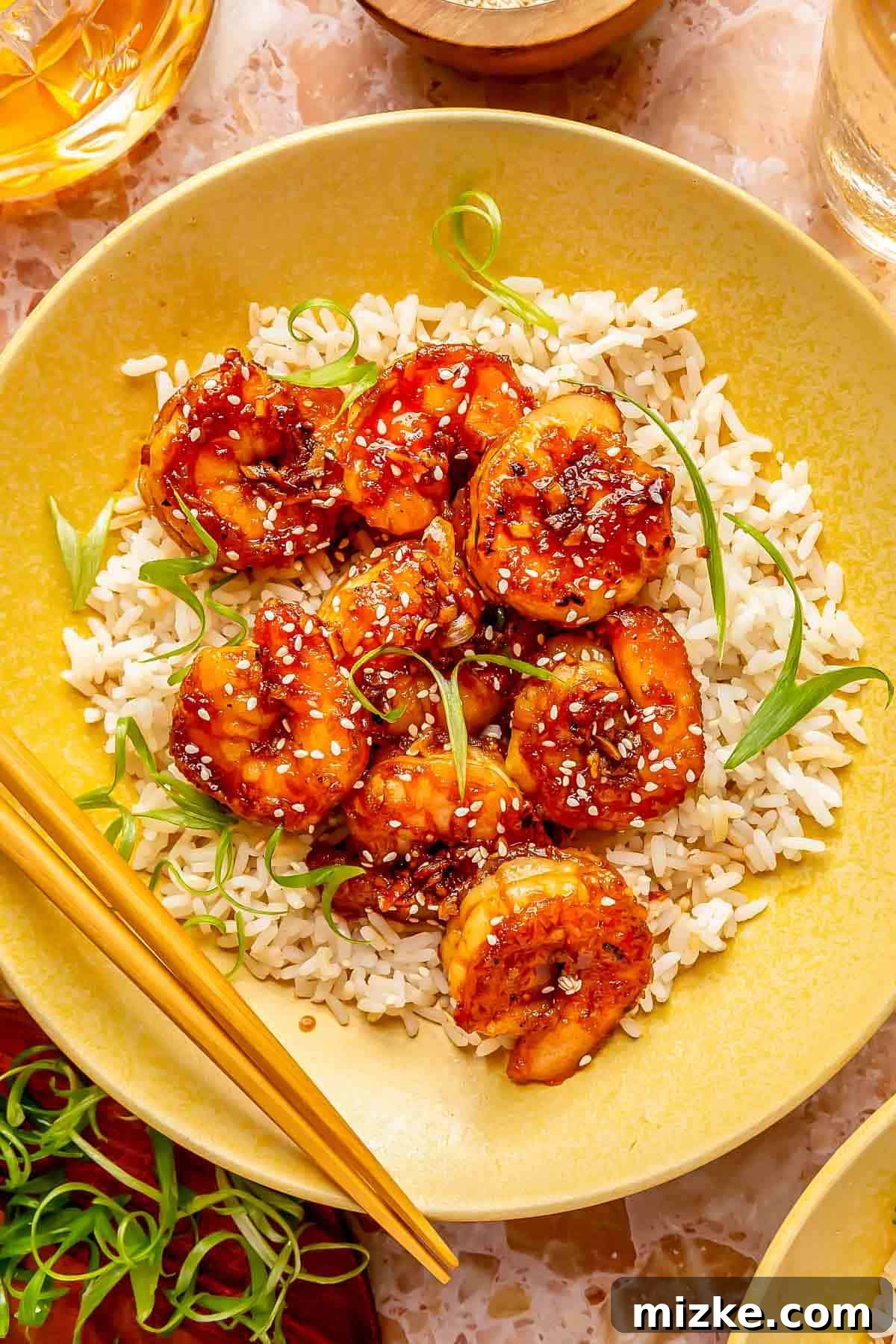 Glazed Honey Garlic Shrimp Stir Fry 3 Honey garlic shrimp served on a bed of fluffy white rice in a vibrant yellow plate, accompanied by wooden chopsticks and garnished with fresh green onions and white sesame seeds for an appetizing presentation.