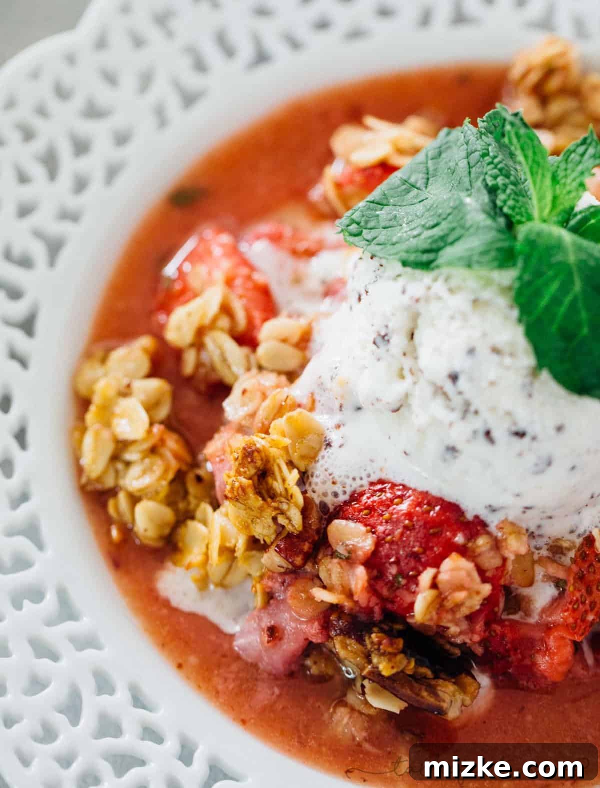 A serving of Strawberry Mint Crisp, perfectly plated with a generous scoop of vanilla ice cream melting slightly on top. The crisp shows off its rich, golden topping and vibrant fruit filling, ready for indulgence.
