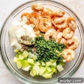 A clear bowl showing a mixture of cooked shrimp, mayonnaise, black pepper, finely chopped celery, and fresh chives, ready to be tossed.