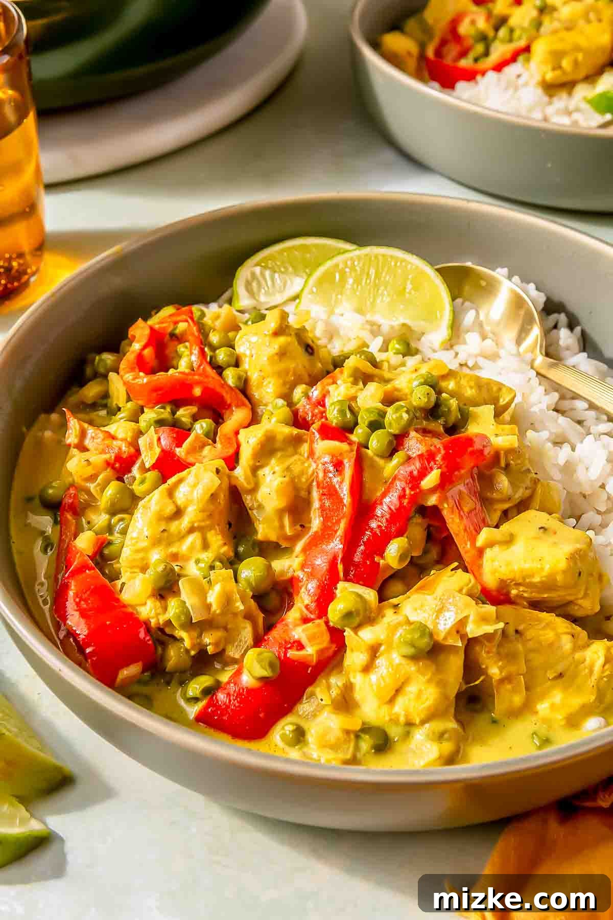 Spicy Thai Green Curry Chicken Rice Bowl 4 A bowl of green curry with chicken, red bell peppers, peas, and white rice, garnished with lime slices, beside a glass of iced tea.