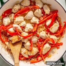 Spicy Thai Green Curry Chicken Rice Bowl 8 A skillet containing cooked chicken pieces with sliced red bell peppers and onions. A wooden spoon rests in the pan. A small bowl of peas is visible in the background.