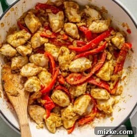 Spicy Thai Green Curry Chicken Rice Bowl 9 A dish of chicken pieces mixed with green curry and red bell peppers cooked in a pot with a wooden spoon. Peas in a small bowl are visible in the background.