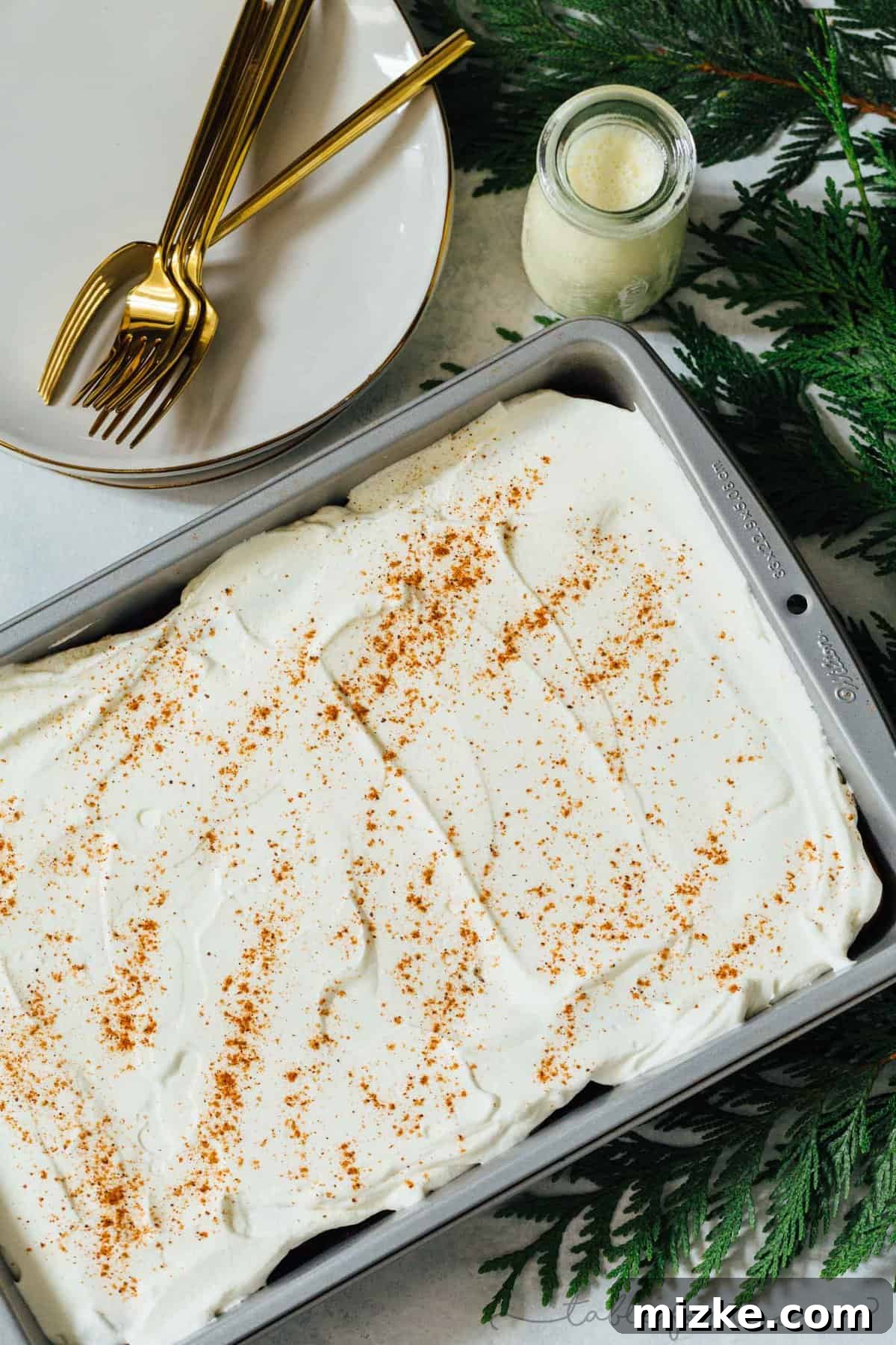 Holiday Eggnog Tres Leches 3 Eggnog tres leches cake in the pan next to festive pine leaves and plates with gold forks, ready for a holiday celebration.