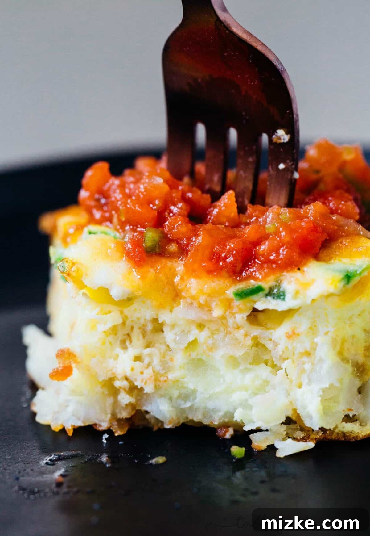 This one-pan cheddar jalapeno tater tot frittata recipe puts a fun twist on a potato and egg frittata, baked inside a crispy tater tot crust. A fork stuck into a slice of cheddar jalapeño tater tot frittata on a black plate.